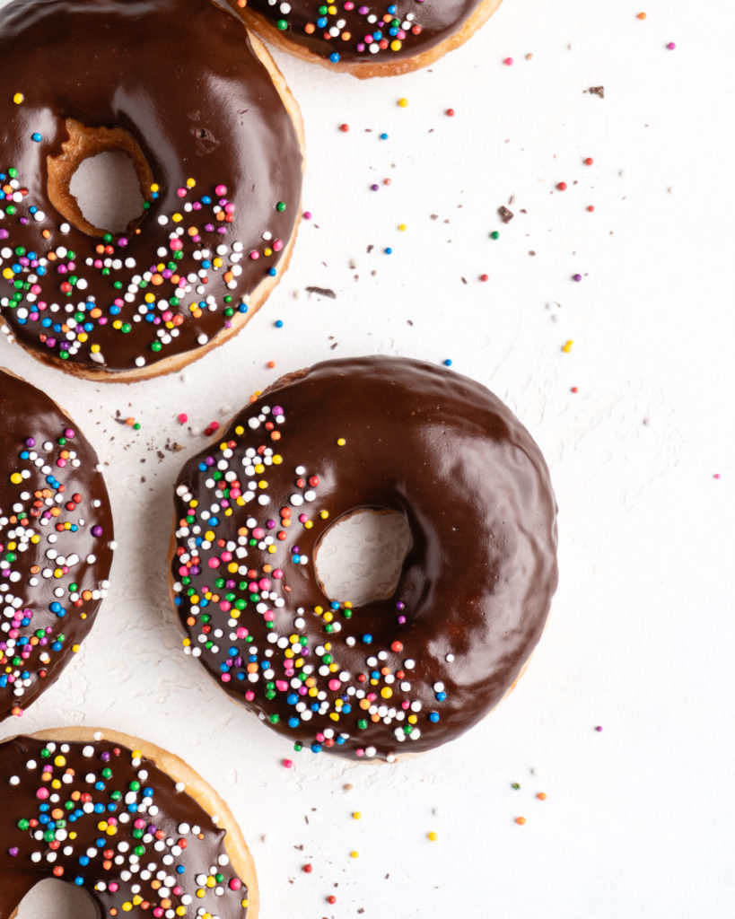 Fluffy golden fried yeast donuts are glazed with a shiny chocolate glaze and colorful round sprinkles