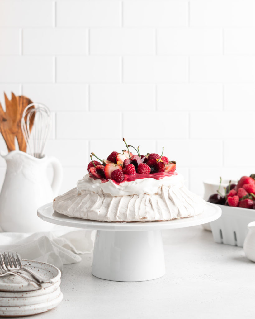 Fluffy and delicious Pavlova is topped with raspberry curd, fresh whipped cream, and loads of summer berries.