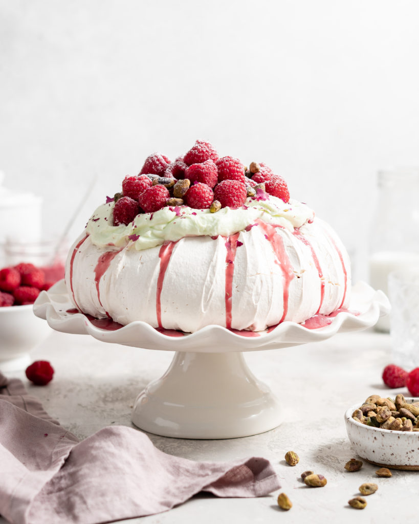 Thick fluffy pavlova topped with whipped pistachio white chocolate ganache and fresh raspberry curd